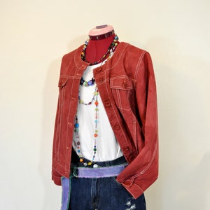 May include: A red jacket with white stitching, worn over a white t-shirt and denim shorts. The outfit is accessorized with beaded necklaces and a purple belt. The shorts have distressed details.