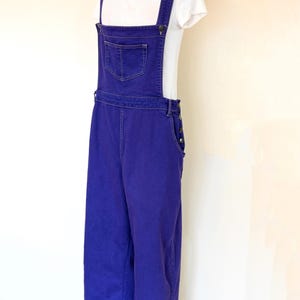May include: Purple denim overalls with a white t-shirt underneath. The overalls have a single front pocket and adjustable straps.