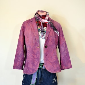 May include: A purple blazer with a tie-dye pattern, paired with a white shirt, denim shorts, and a colorful scarf. The outfit is accessorized with a beaded necklace and a green and purple belt. The blazer has a single button closure.