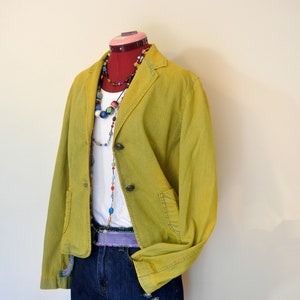 May include: A lime green blazer with two buttons, paired with a white top, denim shorts, and colorful beaded necklaces. The outfit is displayed on a mannequin with a red neck. The shorts have distressed details.