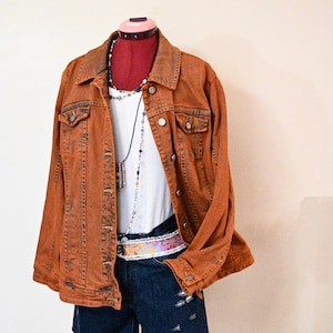 Könnte beinhalten: Eine rostfarbene Jeansjacke über einem weißen gerippten Tanktop und dunkelblauen Jeansshorts mit Used-Optik. Das Outfit wird durch eine mehrfarbige Perlenkette und einen Gürtel mit Blumenmuster in Rosa- und Orangetönen ergänzt.