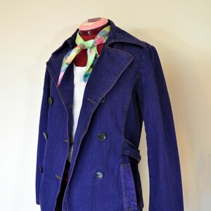 May include: A purple denim jacket with a double breasted closure and a tie-dye scarf. The jacket has a belt and two pockets.