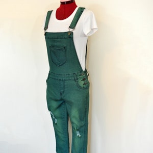 Op de afbeelding: Groene denim overall met distressed details, getoond op een mannequin. De overall heeft verstelbare bandjes, een voorzak en knoopsluitingen. Een wit t-shirt wordt eronder gedragen. Het algehele ontwerp is effen groen.