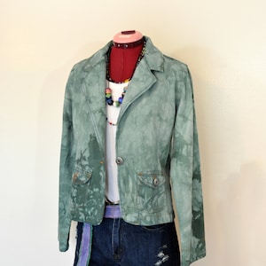 Op de afbeelding: Een groene tie-dye blazer met twee voorzakken, gecombineerd met een spijkerbroek. De outfit bevat een witte top, een kleurrijke kralenketting en een paarse riem. De blazer heeft een enkele knoopsluiting en wordt op een mannequin gepresenteerd.