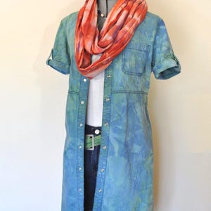 Könnte beinhalten: Ein blaues Button-Down-Hemdkleid mit Batikmuster und hochgekrempelten Ärmeln, kombiniert mit Jeansshorts und einem grünen Gürtel. Ein orange-roter Schal mit Batikmuster ist um den Hals drapiert. Das Outfit wird auf einer Schaufensterpuppe präsentiert.
