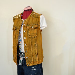 Puede incluir: Chaleco vaquero amarillo mostaza con tachuelas plateadas en los hombros. El chaleco está abotonado y se lleva sobre una camiseta blanca y unos pantalones cortos vaqueros. Un collar de cuentas de colores añade un toque de color.