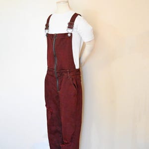 May include: Burgundy denim overalls with a front zipper and distressed knee details. The overalls have adjustable straps and are paired with a white t-shirt. The garment is displayed on a mannequin.