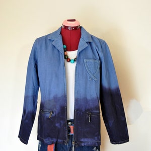 May include: A blue denim jacket with a tie-dye effect, fading from light blue to dark blue. The jacket has a zipper closure, two pockets on the front, and a small pocket on the left chest. The jacket is worn over a white t-shirt and blue denim shorts.