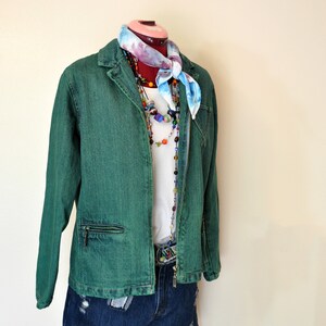 May include: A green denim jacket with a zipper closure, paired with denim shorts. The outfit includes a white t-shirt, colorful beaded necklaces, and a patterned scarf. The mannequin is on a red stand.