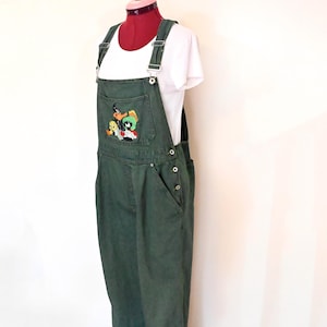 May include: Dark olive green denim overalls featuring a chest pocket with an embroidered graphic of Looney Tunes characters including Tweety Bird and Marvin the Martian. The overalls have adjustable metal buckles on the shoulder straps and side buttons.