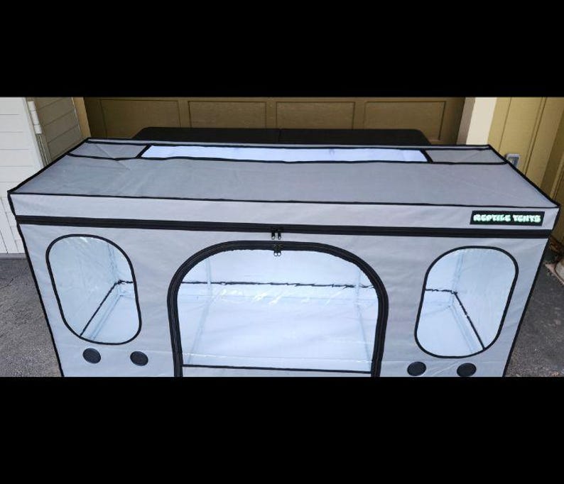 May include: A gray reptile enclosure with black trim and clear viewing windows. The enclosure has a zippered front door and the text "REPTILE TENTS" on the side. The top has a rectangular window.