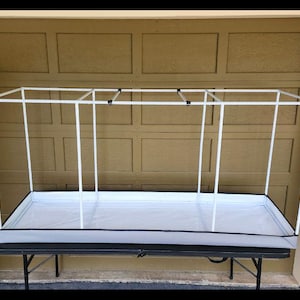 May include: A white PVC frame greenhouse structure with a rectangular base and a white interior. The frame is supported by vertical poles and connected at the top. The structure sits on a black folding table.
