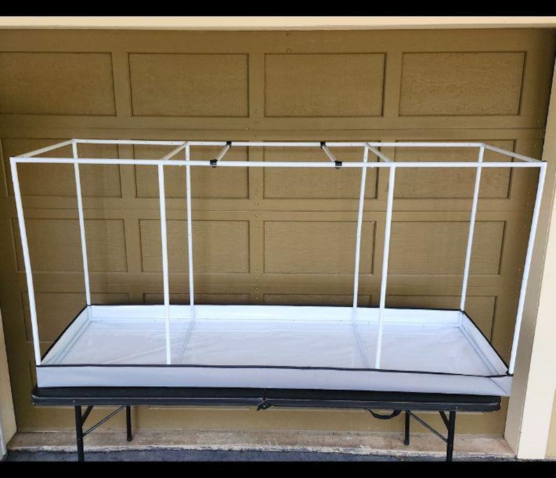 May include: A white PVC frame greenhouse structure with a rectangular base and a white interior. The frame is supported by vertical poles and connected at the top. The structure sits on a black folding table.