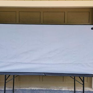 May include: A large, light gray screen stretched across a black folding table. The screen is rectangular with black trim and secured with black fasteners. The table has black metal legs.