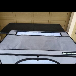 May include: A gray reptile enclosure with a black zipper and the text "REPTILE TENTS" in a green font. The enclosure has a white panel and is placed on a black table.