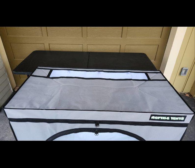 May include: A gray reptile enclosure with a black zipper and the text "REPTILE TENTS" in a green font. The enclosure has a white panel and is placed on a black table.