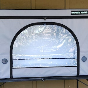 May include: A reptile enclosure with a clear front panel and a gray exterior. The enclosure has a black zipper and trim, and the text "REPTILE TENTS" is visible on the top. It features ventilation holes and a black base.