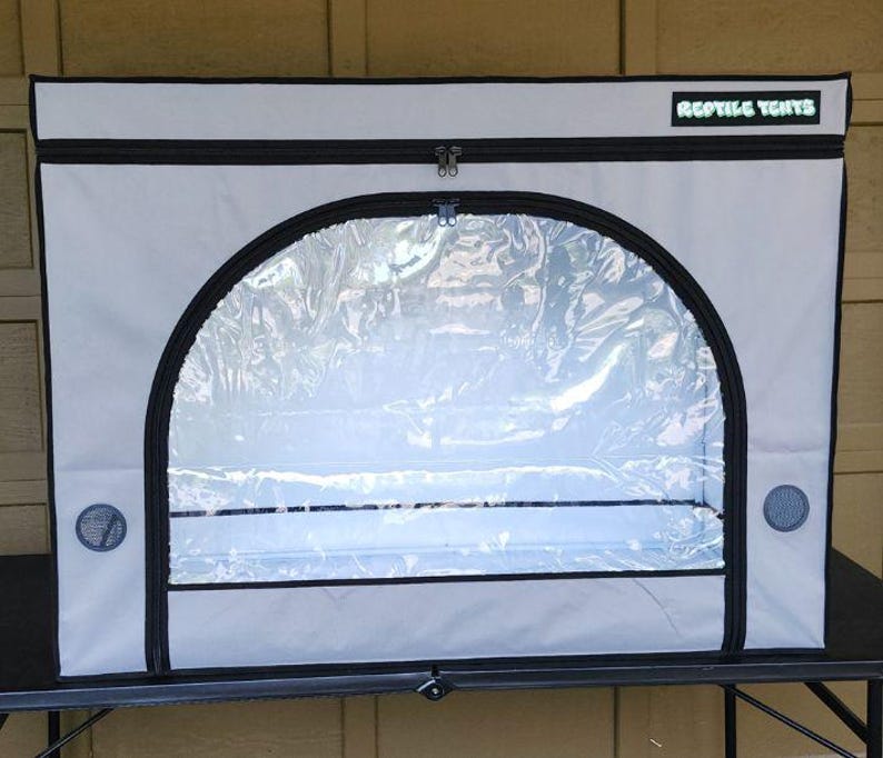 May include: A reptile enclosure with a clear front panel and a gray exterior. The enclosure has a black zipper and trim, and the text "REPTILE TENTS" is visible on the top. It features ventilation holes and a black base.