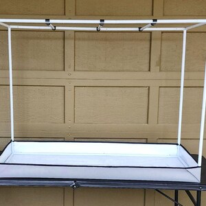 May include: A white rectangular plant stand with a black trim and a white frame. The stand has a rectangular base and a tall, open frame. The stand is designed to hold plants and is placed on a black table.