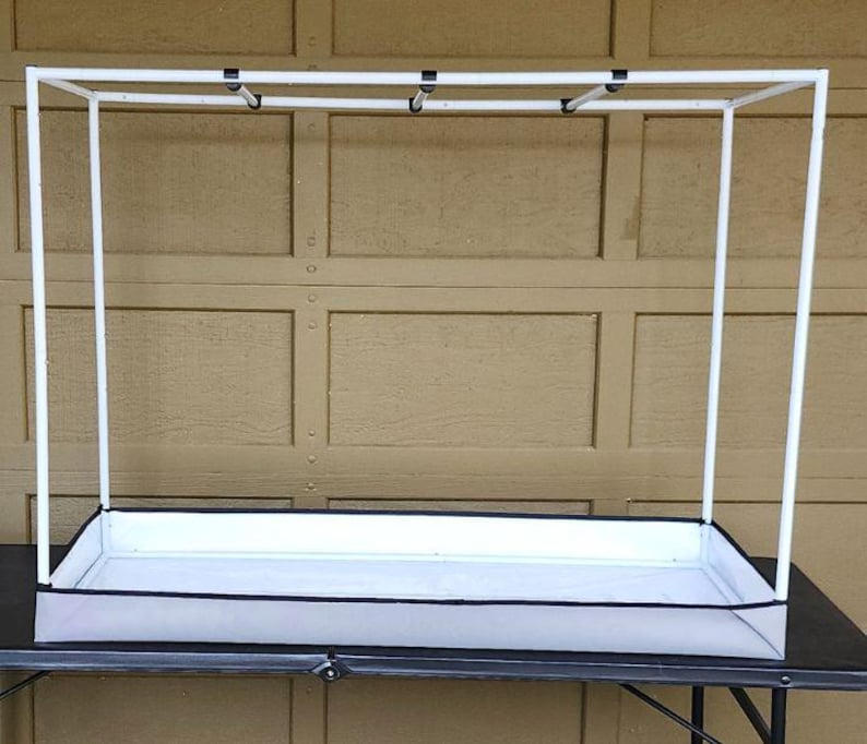 May include: A white rectangular plant stand with a black trim and a white frame. The stand has a rectangular base and a tall, open frame. The stand is designed to hold plants and is placed on a black table.
