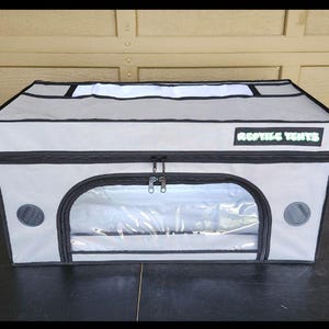May include: A gray reptile enclosure with a black zipper and a clear front window. The top of the enclosure has a white panel and the words "REPTILE TENTS" in green. There are two circular vents on the sides.