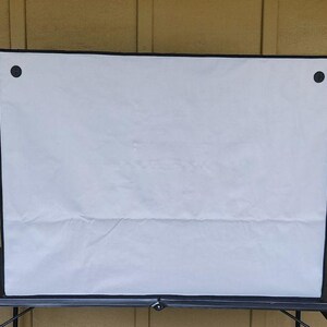 May include: A white rectangular cover with a black border and two black fasteners. The cover is made of a soft, possibly fabric material, and is placed on a black table. The background is a light brown wooden wall.