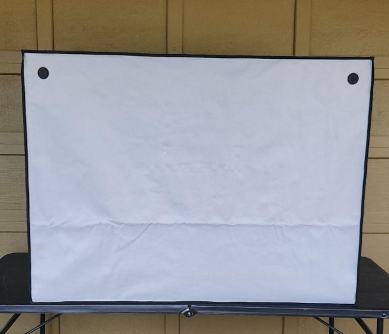 May include: A white rectangular cover with a black border and two black fasteners. The cover is made of a soft, possibly fabric material, and is placed on a black table. The background is a light brown wooden wall.