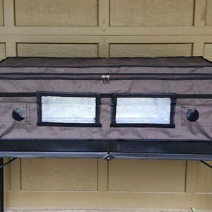 May include: A brown and black grow tent with two clear viewing windows and two circular ventilation ports. The tent has a zippered top and is sitting on a black table. The tent is designed for indoor gardening.
