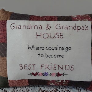 May include: A patchwork pillow with a white front panel featuring the embroidered text "Grandma & Grandpa's HOUSE Where cousins go to become BEST FRIENDS" with a small floral design at the bottom.