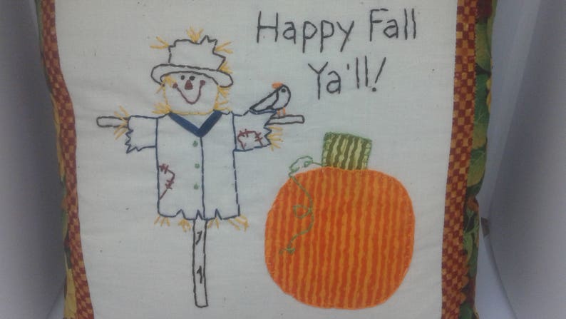 May include: Embroidered pillow with a scarecrow, pumpkin, and bird. The pillow has a white background with a red and green border. The text "Happy Fall Y'all!" is embroidered on the pillow.