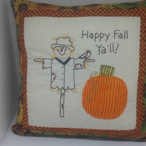 May include: A white pillow with a brown, green, and yellow border. The pillow has an embroidered image of a scarecrow with a pumpkin and the text "Happy Fall Ya'll!"