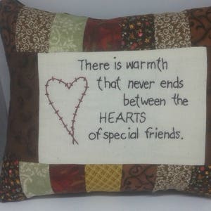 May include: A decorative pillow with a patchwork design in shades of brown, green, and yellow. The center features a white fabric with embroidered text and a heart that reads, "There is warmth that never ends between the HEARTS of special friends."