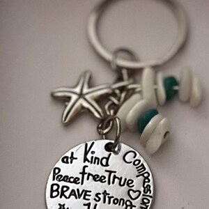 May include: Silver keychain with a round charm that says "at Kind Compassionate Peace free True BRAVE strong Happy thankful". The charm is attached to a silver starfish, a white seashell, and a turquoise bead.