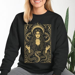 May include: Black sweatshirt featuring a gold illustration of a woman with long hair, a sun halo, and snakes. The design has a vintage, mystical aesthetic with intricate details and a decorative border. The artwork is centered on the front of the sweatshirt.