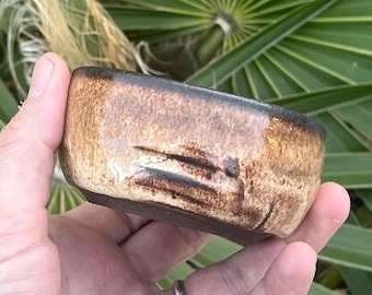 May include: A small, rectangular ceramic bowl with a textured, brown and tan exterior and a dark brown rim. The bowl has a dark brown base and is held in a hand against a backdrop of green palm leaves.