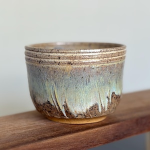 May include: A handmade ceramic bowl with a textured, layered rim. The bowl features a blend of earthy tones, including brown, beige, and hints of blue and green. The glaze has a dripped effect, adding to its unique, artisanal appearance.