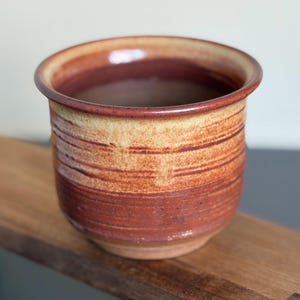May include: A handmade ceramic cup with a rounded base and a slightly flared rim. The cup features horizontal bands of reddish-brown and tan hues, creating a rustic, earthy aesthetic. The cup is likely used for drinking beverages.