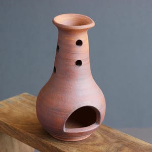 May include: A terracotta oil burner with a bulbous base, a flared rim, and a large arched opening at the bottom. The upper neck features several small circular ventilation holes arranged vertically, showcasing a natural matte clay finish.
