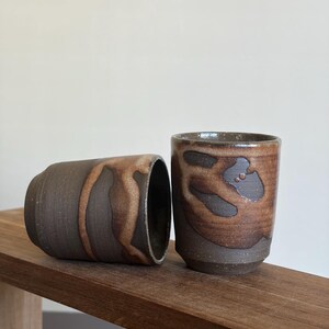 May include: Two handmade ceramic cups with a dark brown base and a lighter brown, organic pattern. One cup is upright, the other is on its side. The cups are on a wooden surface.