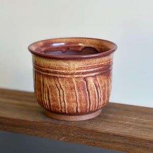 May include: A handmade ceramic planter with a textured, vertical striped design. The planter features a warm color palette of brown, tan, and reddish-brown hues. The rim and interior are a darker shade of brown.
