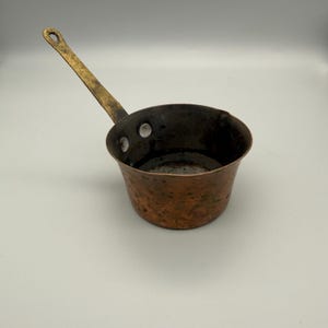 May include: A small, round copper saucepan with a hammered exterior finish and a dark, weathered patina. It features a long, angled brass handle attached to the side with two visible rivets. The interior is dark and shows signs of heavy use.