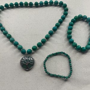 May include: A jewelry set featuring a turquoise beaded necklace with a silver tree of life pendant, and two matching bracelets. The beads are a vibrant teal color, and the set is displayed on a neutral fabric.