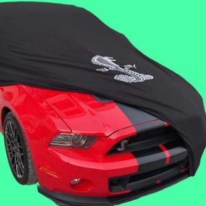 May include: A black car cover with a white embroidered snake logo, covering a red Ford Mustang with black racing stripes.
