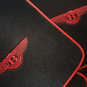 Könnte beinhalten: Nahaufnahme einer schwarzen, strukturierten Oberfläche mit zwei roten geflügelten Emblemen und einem rot genähten Rand. Die Embleme zeigen ein stilisiertes "B" in einem Kreis mit Flügeln. Das Design ist elegant und modern.