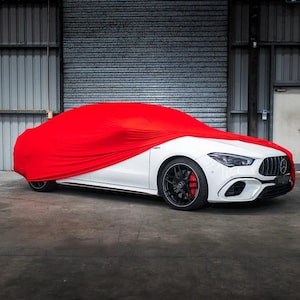 May include: A white sports car with black rims and red brake calipers is partially covered by a bright red car cover. The car is parked inside a garage with a gray concrete floor and metal walls.