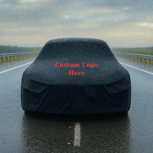 May include: A black car cover on a wet road with the text "Custom Logo Here" in red. The car cover is draped over a vehicle, obscuring its shape. The background shows a rainy sky and a highway.