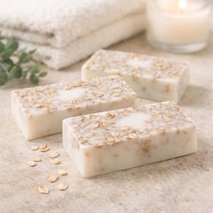 Puede incluir: Tres barras rectangulares de jabón artesanal con copos de avena incrustados. El jabón es de color blanco cremoso, con un ligero tono beige de la avena. Una pila de toallas blancas y una vela encendida están en el fondo.