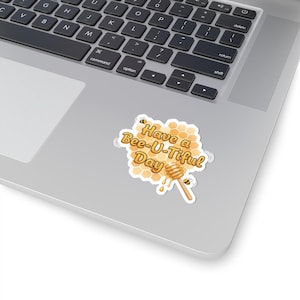 May include: A sticker with the phrase "Have a Bee-U-tiful Day" in yellow and brown, featuring a honeycomb design, a honey dipper, and two cartoon bees. The sticker is placed on a laptop.