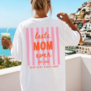 May include: White t-shirt with a pink and orange graphic on the back. The graphic reads "beste MOM ever" with pink vertical stripes and the text "EST. 2017" and "DEIN TEXT STEHT HIER" below. The person is holding a green drink.
