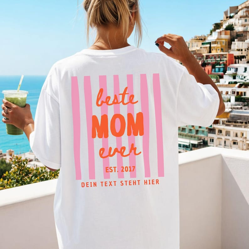 May include: White t-shirt with a pink and orange graphic on the back. The graphic reads "beste MOM ever" with pink vertical stripes and the text "EST. 2017" and "DEIN TEXT STEHT HIER" below. The person is holding a green drink.